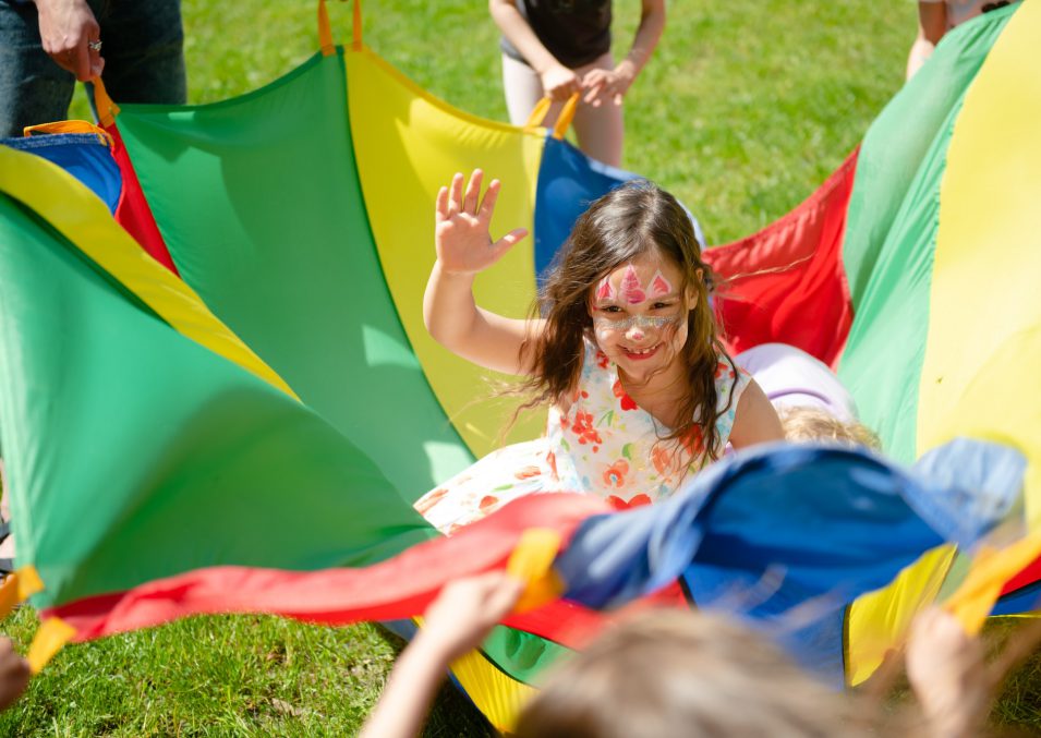 Girl with water make up on her face having fun in multicoloured play parachute on kid’s party.
